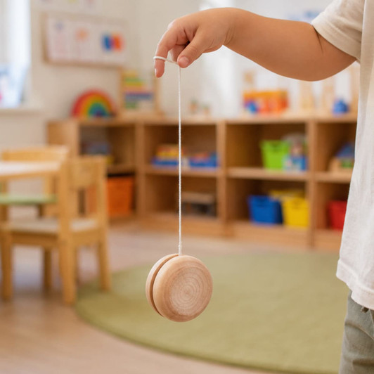 A child's hand holds a wooden yo-yo with a natural finish, dangling on a string in a bright, playful indoor space.