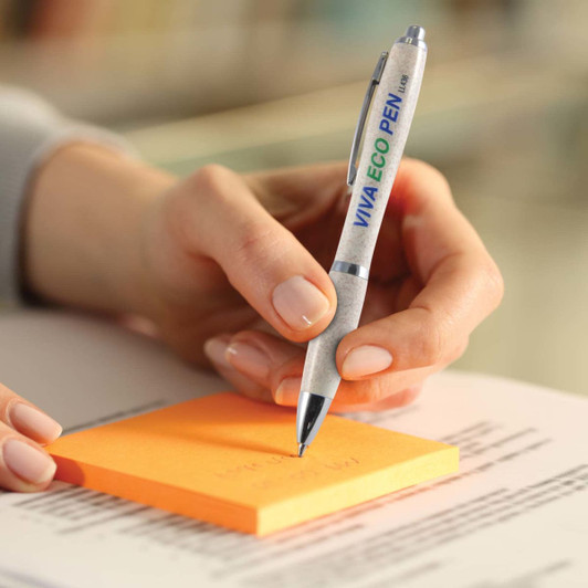 A hand holds a silver and green pen, writing on an orange sticky note with printed text below. The pen features a logo.