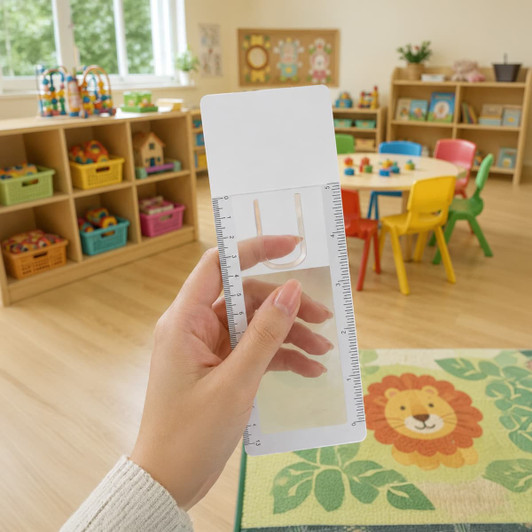 A clear bookmark magnifier ruler held in a hand, set against a colourful classroom with wooden shelves and toys.