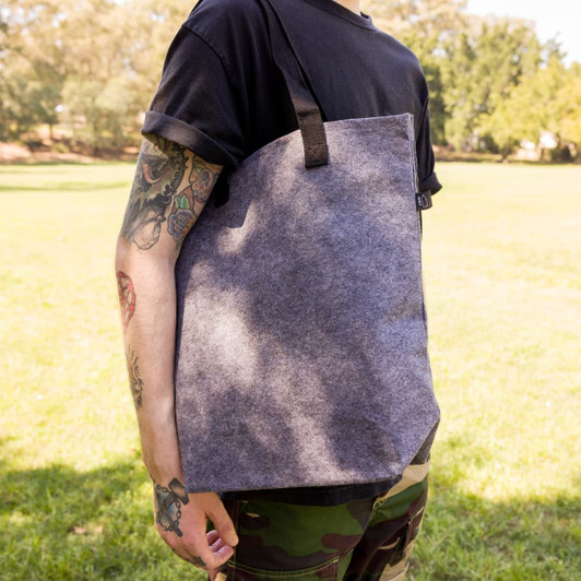 A grey felt tote bag with black straps, held by a person in a casual outfit, set against a park background.