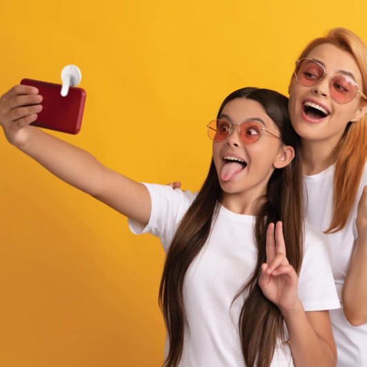 A selfie is being taken by two girls wearing sunglasses and white t-shirts, with a bright yellow background.