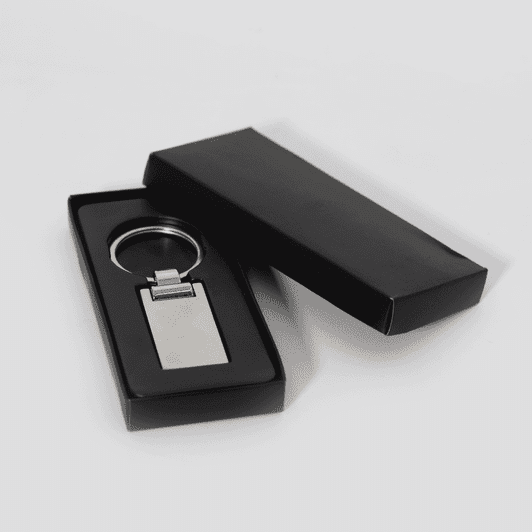 A silver keychain displayed inside a black box. The keychain has a rectangular shape with a polished finish.