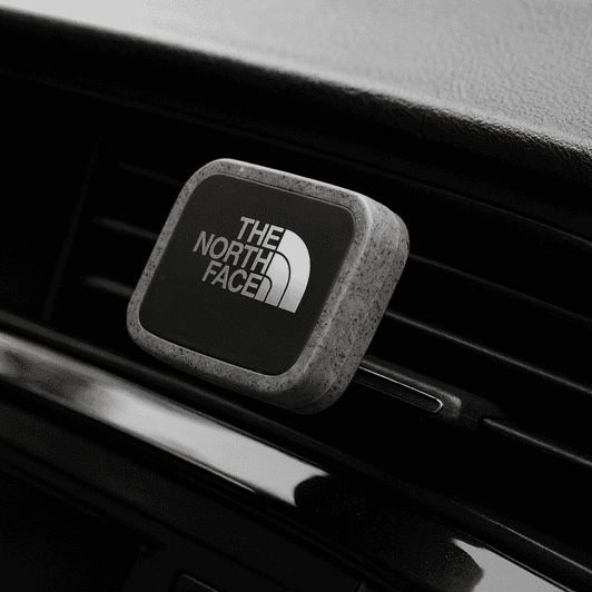 A rectangular car vent mount in silver with a logo, designed for holding phones, against a dark background.
