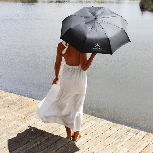 A black umbrella with a logo held by a person in a white dress, standing by the water.
