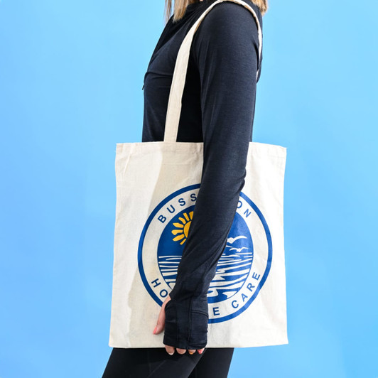 A cream-coloured tote bag with a logo, held by a person against a blue background.
