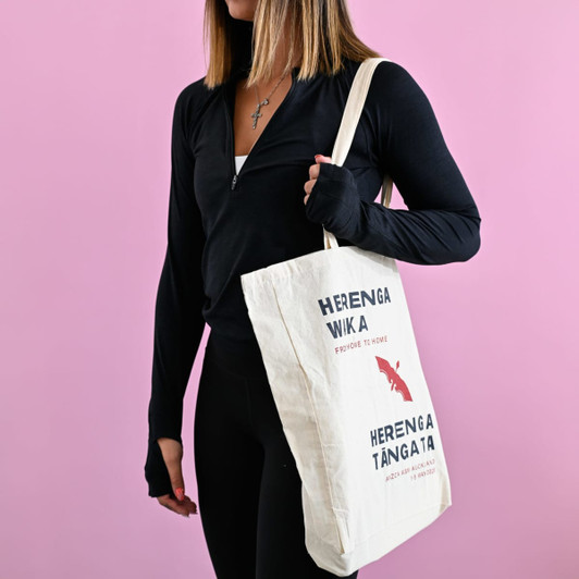 A cream-coloured tote bag with a gusset, featuring a logo and text in red and blue, held by a person in black attire.