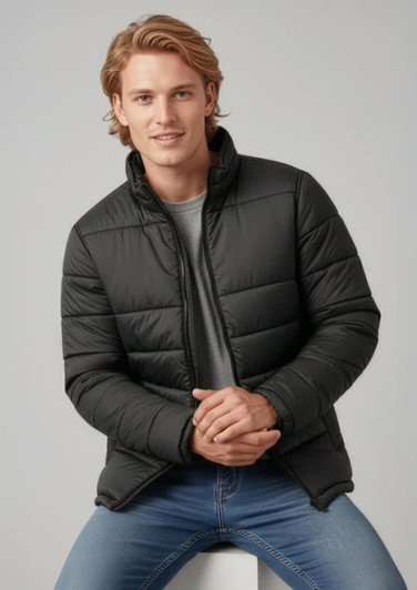A man wearing a black puffer jacket, sitting on a white surface, with light jeans and a grey t-shirt.