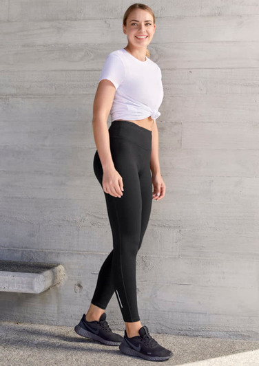A woman wearing black flex leggings and a white tied T-shirt, standing against a textured wall. The leggings have a logo.