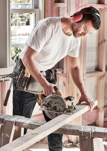 A man in a white T-shirt uses a circular saw on a wooden plank, wearing safety glasses and noise-cancelling headphones.