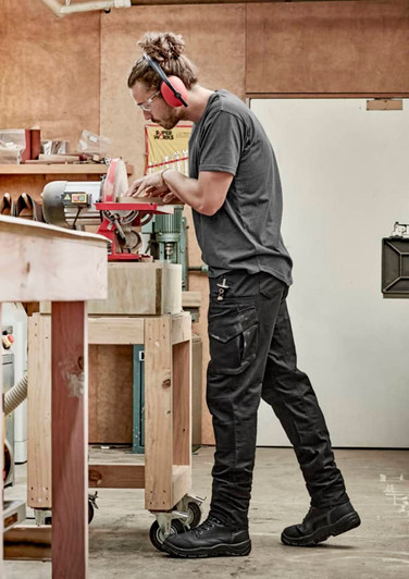 A person wearing black rugged cooling stretch pants stands in a workshop, using a tool. The pants feature pockets and have a logo.