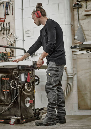 A man wearing black work pants stands by a workshop table, handling tools in a well-equipped space.