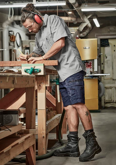 A man wearing grey work shirt and navy shorts stands at a workbench, using a pencil to mark wood. He has ear protection on.