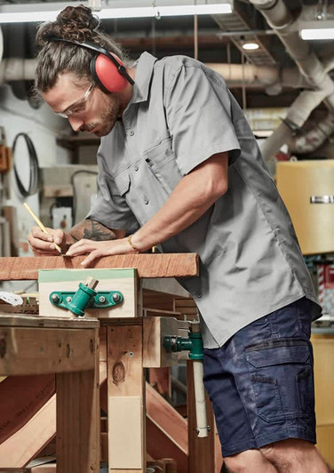 A man wears a grey short sleeve shirt with a logo, using a pencil at a workbench in a workshop setting.