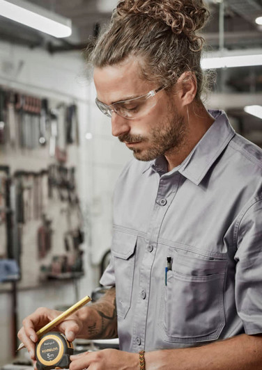 A man wearing a grey short sleeve work shirt, safety glasses, examining a measuring tape in a workshop setting.