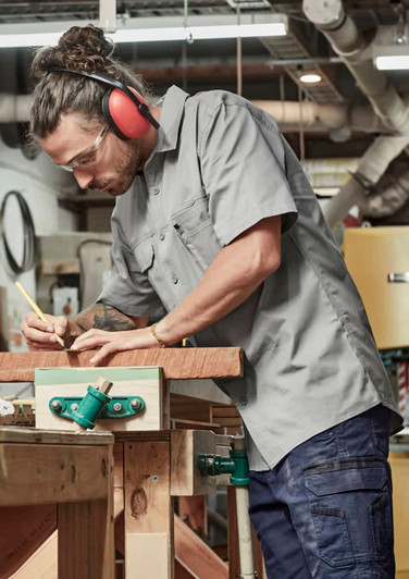 A lightweight grey short sleeve work shirt worn by a man using a pencil on wood in a workshop. The shirt has a logo.
