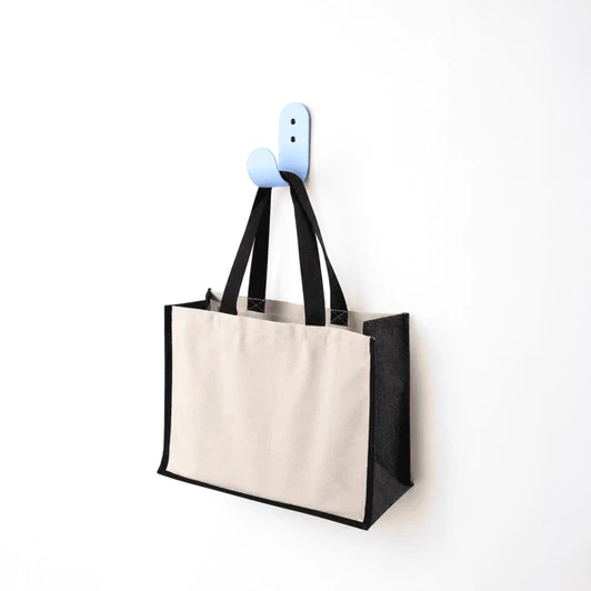 A functional tote bag in beige and black hangs on a blue hook against a white wall. The bag has handles and a logo.