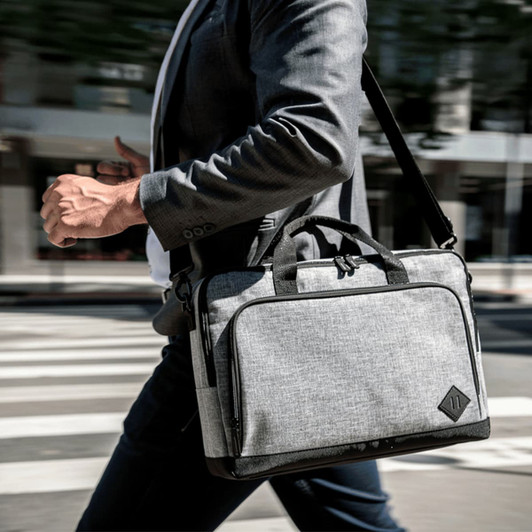 A grey computer briefcase with a shoulder strap and handles, featuring a logo, carried by a person in a suit.