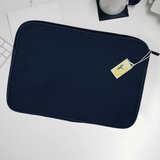 A navy recycled canvas laptop sleeve with a zipper and a tag, resting on a white desk alongside a keyboard and mug.