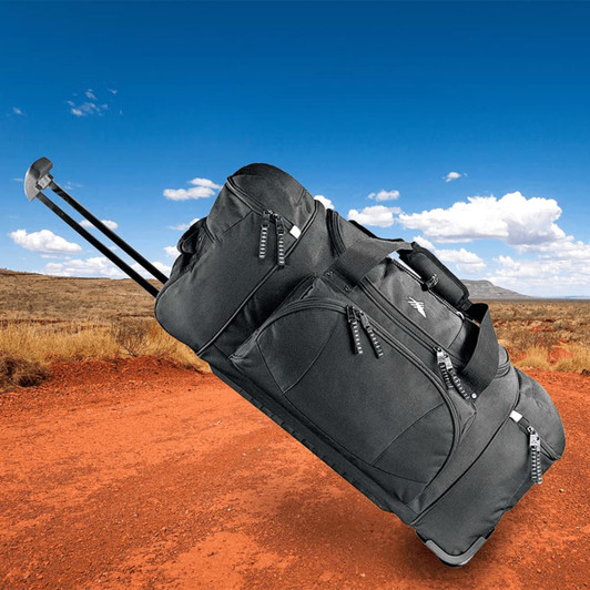 A wheeled duffel bag in black with multiple zippers, positioned on a dirt path against a blue sky.