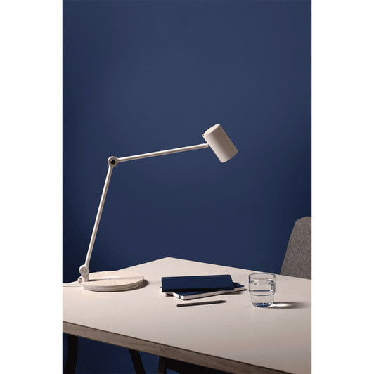 A white desk lamp with an adjustable arm, a blue notebook, and a glass of water on a light wood table against a blue wall.