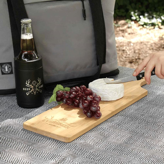A bamboo serving board with cheese and grapes, alongside a drink bottle, set on a textured surface outdoors.