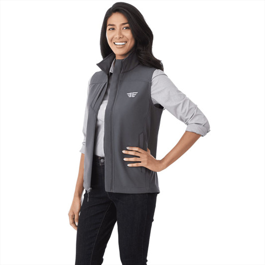 A woman models a grey Stinson Softshell Vest, featuring a logo, paired with a light shirt and dark jeans.