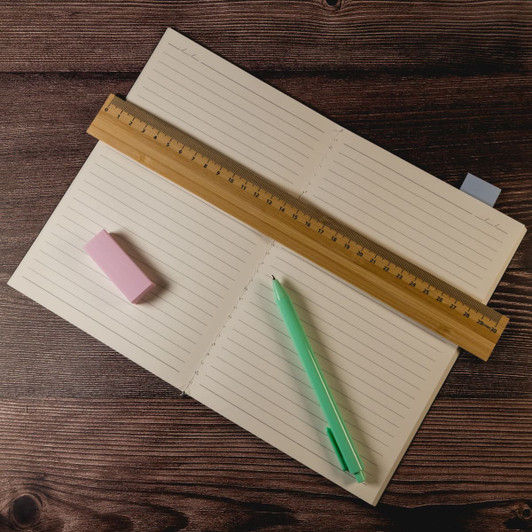 A bamboo 30cm ruler lies on an open lined notebook, accompanied by a pink eraser and a green pen.
