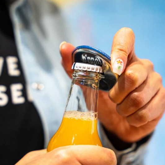 A drink bottle opener with a blue handle is being used to cap a glass bottle of yellow beverage.
