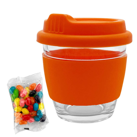 A glass coffee cup with an orange silicone lid and band, accompanied by a bag of colourful jelly beans.