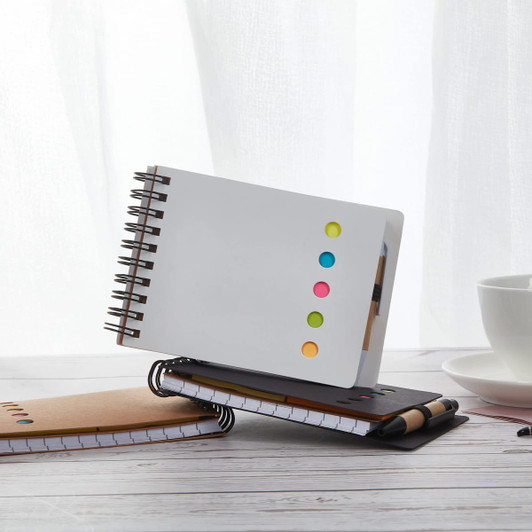 A white spiral-bound note pad with colourful sticky dots, placed on a wooden table alongside two other notebooks.