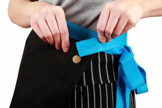 A black canvas short waist apron with a bright blue tie, being fastened around the waist.
