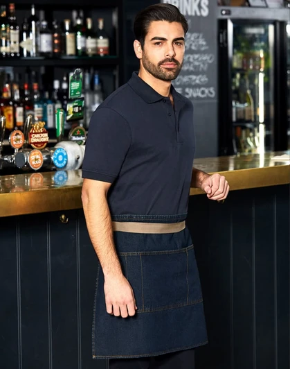 A man wearing a denim half apron stands at a bar, featuring a logo on the apron and a dark background with shelves of drinks.