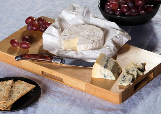 A cheese board set featuring a wooden board, a knife, various cheeses, crackers, grapes, and a bowl of grapes.