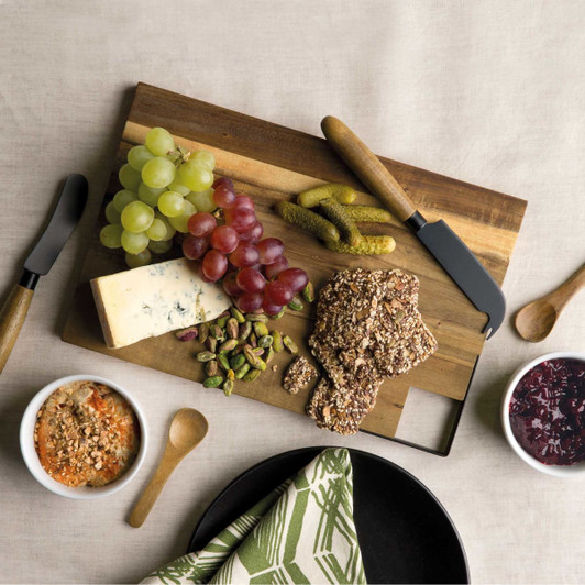 A wooden cheese board displaying grapes, olives, nuts, and crackers, accompanied by cheese knives and small bowls.