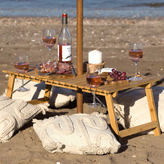 A bamboo table with wine, glasses, and food, set on a sandy beach with decorative pillows.