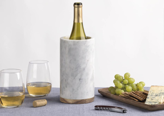 A drink bottle sits in a white marble cooler, accompanied by two glasses of wine, grapes, crackers, and cheese.
