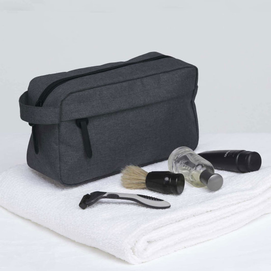 A dark grey toiletry bag with grooming items including a razor, shaving brush, and travel-sized bottles, placed on a white towel.