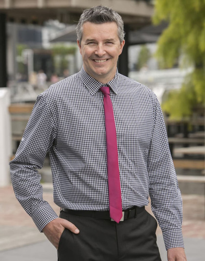 A man in a blue and white gingham check long-sleeve button-up shirt, wearing a pink tie and black trousers.