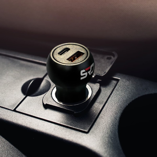 A black USB car charger with two ports, featuring a round design and a logo, set in a vehicle's dashboard.