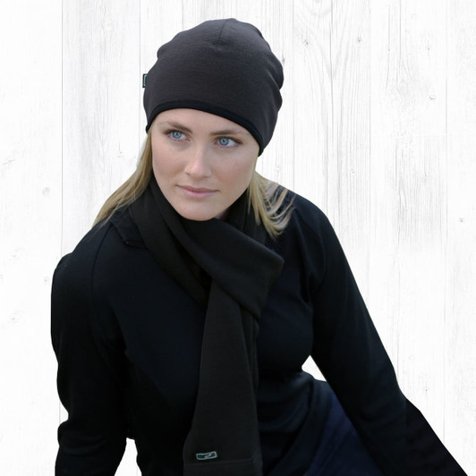 A black Merino Arctic Beanie worn by a woman, paired with a matching scarf, against a light wooden background.