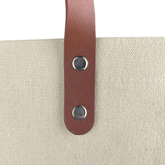 A beige cotton tote bag featuring a brown leather handle with silver metal studs.