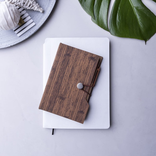 A wooden textured sticky note pad with an elastic closure, placed on top of a white notebook and a grey surface.