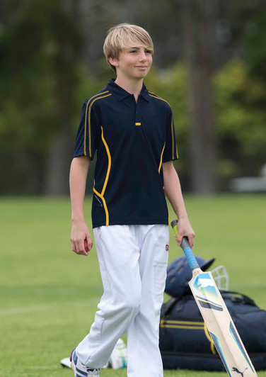 A youth wearing a navy polo shirt with yellow accents, walking on a cricket field with sporting equipment.