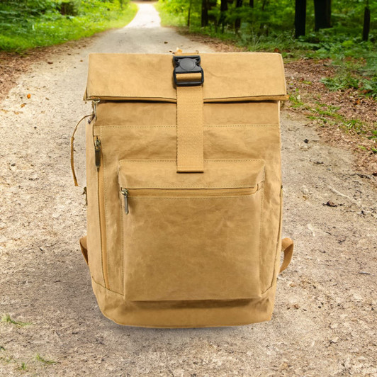 A tan kraft paper laptop backpack with a front pocket, positioned on a dirt path surrounded by greenery.
