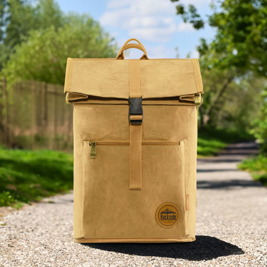 A yellow kraft paper laptop backpack with a fold-over top and front zip pocket, set against a scenic outdoor path.