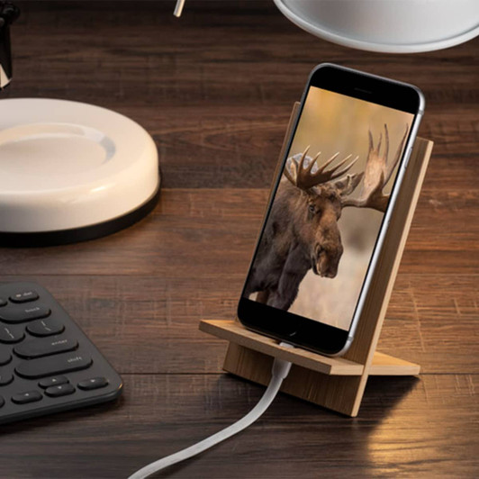 A bamboo phone holder displaying a phone with an image of a moose, set on a wooden desk beside a keyboard and lamp.