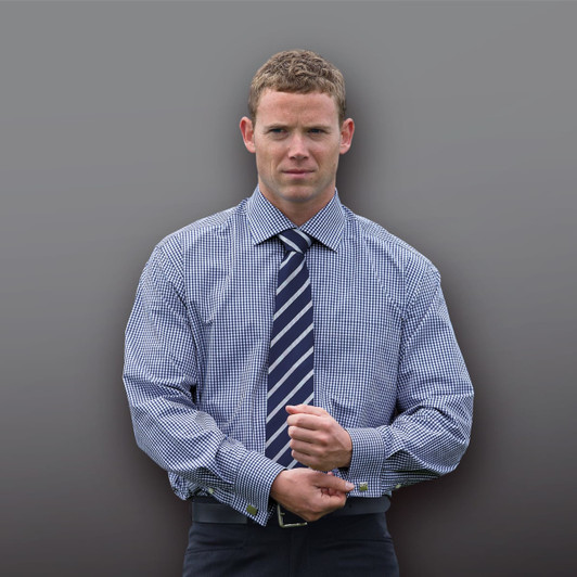 A man in a blue and white checked button-up shirt with a navy tie, posed against a grey background.