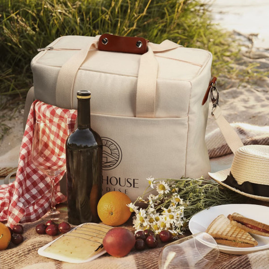 A beige cooler tote bag with a logo, a drink bottle, cheese, fruit, crackers, and a checkered cloth on grass.