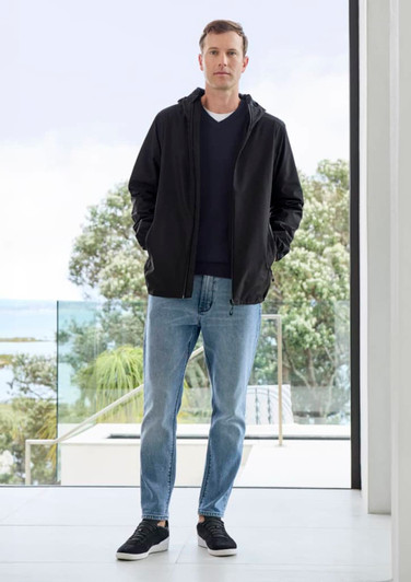 A man wearing a black rain jacket, blue jeans, and black shoes, standing indoors with a view of greenery outside.