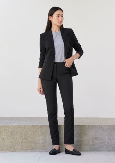 A woman in a black suit with a grey top and black shoes stands against a neutral background. The outfit includes tailored slacks.
